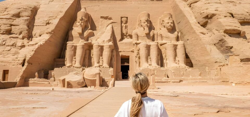 A visitor with a long blonde ponytail and a white shirt stands in front of the Great Temple of Abu Simbel, with its four seated statues of Ramesses II carved into the rock